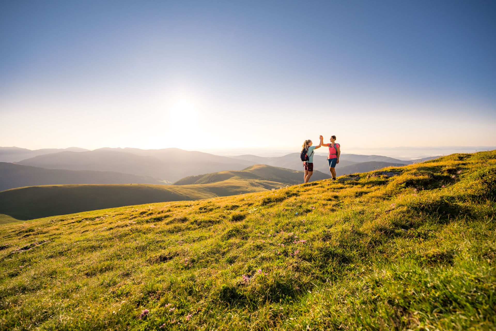 Zwei Wanderer klatschen sich auf einer Almwiese bei Sonnenuntergang ab