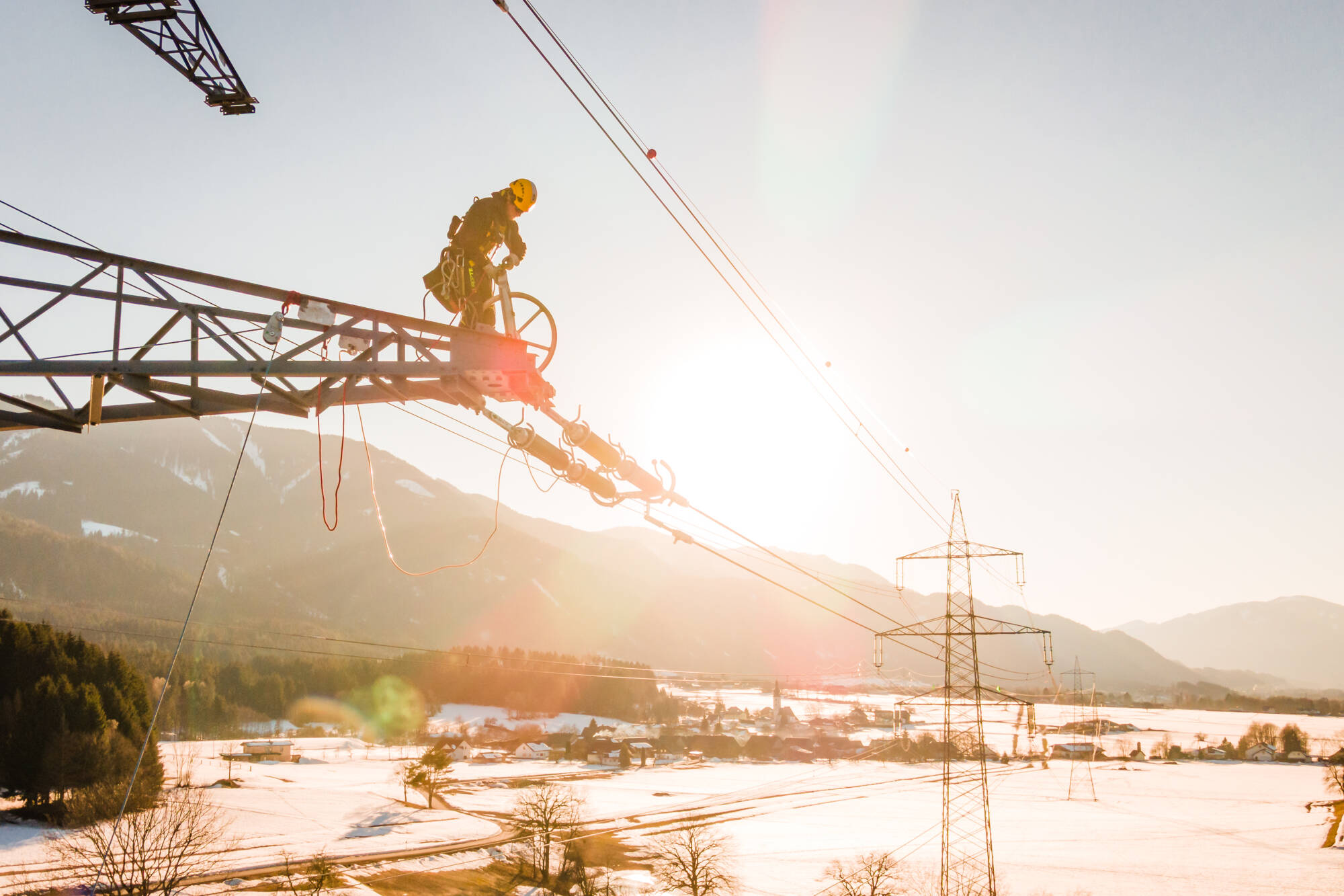 Monteur auf einem Strommast im Gegenlicht, Drohnenaufnahme über einem verschneiten Tal