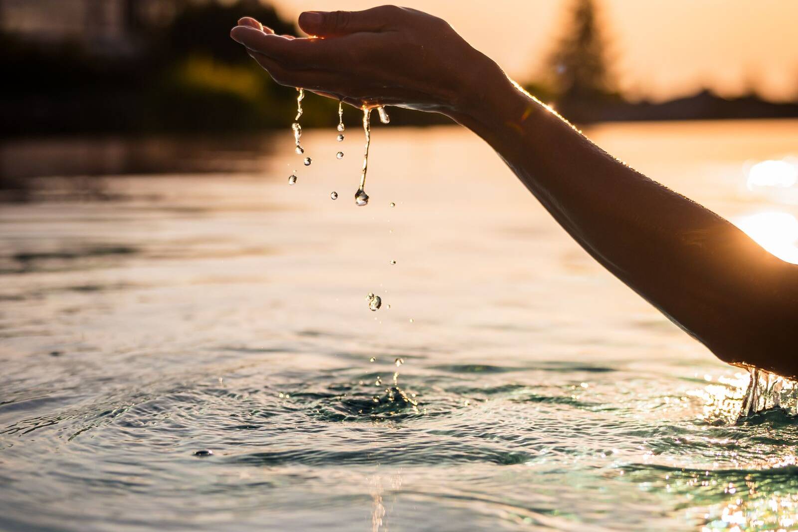 Hand schöpft Wasser im Infinity-Pool des Mountain Resort Feuerberg bei Sonnenaufgang – Wellness- und Hotelfotografie